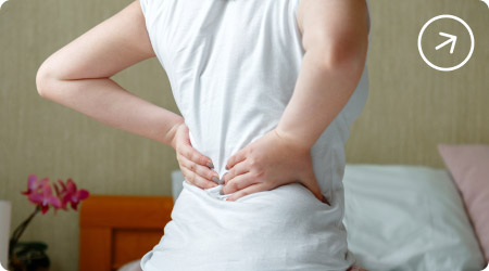 Mujer sentada en su cama sufriendo de dolor lumbar, tocándose la parte baja de la espalda.